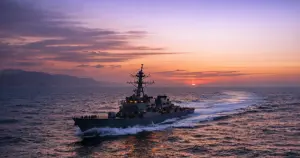 U.S. Navy warship sailing alone through the Strait of Hormuz at dusk with dramatic sky and ocean spray