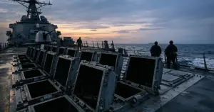 Empty missile launch cells on Navy warship deck at dusk with crew silhouettes working in background