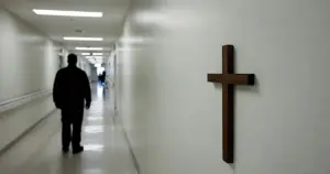 Silhouetted healthcare worker walking down hospital corridor with crucifix on wall